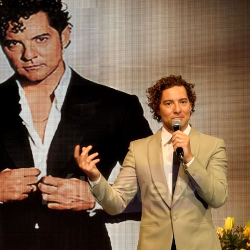Artista en escenario frente a pantalla gigante con su retrato, sosteniendo micrófono.