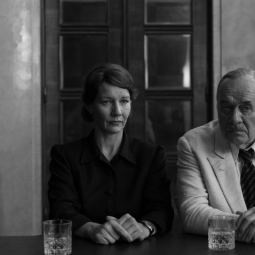 Fotografía en blanco y negro donde la actriz Sandra Hüller y el actor Hanns Zischler permanecen sentados juntos detrás de una mesa de madera con vasos de cristal, vistiendo trajes formales mientras miran hacia el frente en un encuadre contenido.   