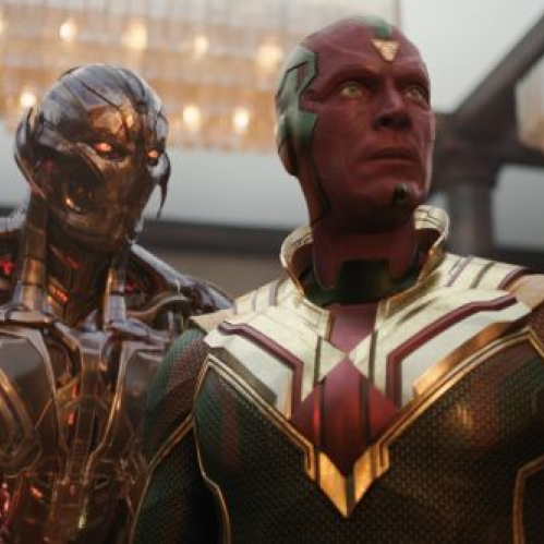 El androide Vision de rostro rojo y traje verde con dorado permanece de pie junto a la figura metálica de Ultron en un salón con candelabros de cristal al fondo.