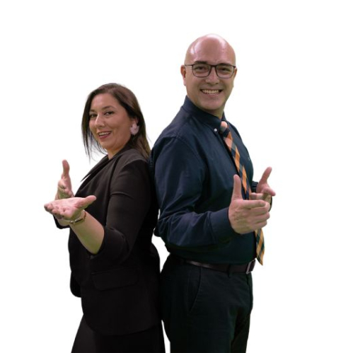 Una mujer vestida con saco negro y un hombre con camisa azul y corbata posan de espaldas juntos con las manos extendidas haciendo un gesto frente a un fondo negro.