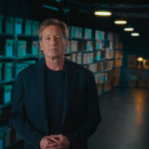 El actor y conductor David Duchovny parado de frente con las manos entrelazadas en primer plano. Viste un saco azul marino sobre una playera oscura. Se encuentra en una habitación iluminada de forma tenue con estanterías de metal repletas de cajas de archivo de cartón y expedientes. La iluminación ambiental resalta con tonos azules y cálidos al fondo.