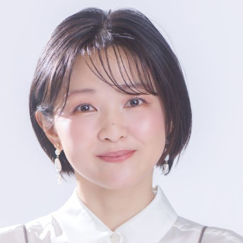 Fotografía de estudio de la actriz de voz japonesa Chiwa Saito. Aparece en plano medio, luciendo cabello corto estilo bob de color castaño oscuro con fleco recto. Viste una blusa blanca de cuello cerrado debajo de un top translúcido en tono gris drapeado. Sonríe mirando hacia la cámara con un fondo claro e iluminado.