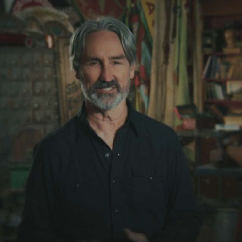 El presentador Mike Wolfe mirando a la cámara con una sonrisa ligera. Viste una camisa de manga larga oscura estilo vaquero sobre un fondo de taller o depósito con herramientas, carteles vintage y estanterías de madera en tonos cálidos.