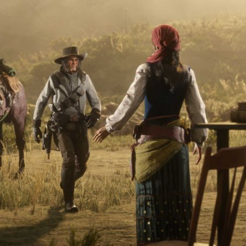 Captura de pantalla de Red Dead Online ambientada al atardecer en un entorno desértico. A la derecha, Madam Nazar aparece de espaldas vistiendo su vestimenta tradicional con pañuelo rojo, falda azul con patrones dorados y faja amarilla, junto a su gramófono. A la izquierda, un vaquero camina hacia ella con sombrero gris, abrigo ligero y bandoleras, acompañado por un caballo marrón cargado con equipaje en segundo plano.