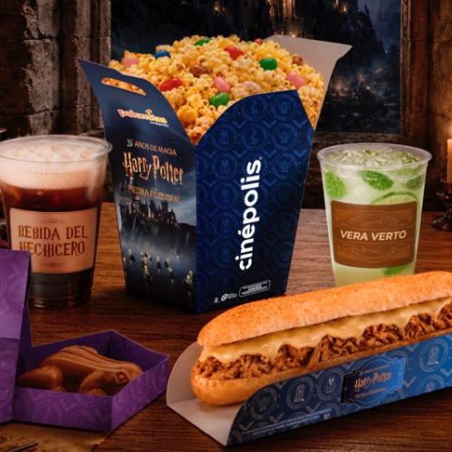 Fotografía en plano medio que muestra la mesa temática de alimentos inspirada en Harry Potter dentro de una ambientación mágica de piedra con velas encendidas al fondo. Al centro destaca una caja azul de palomitas con grageas de colores decorada con el título "Harry Potter y la Piedra Filosofal" y el logotipo de Cinépolis. En primer plano a la derecha se observa un emparedado largo de queso fundido sobre una bandeja azul con motivos dorados. A la izquierda, una caja morada abierta muestra una rana de chocolate. A los lados acompañan los dos vasos con bebidas: a la izquierda la "Bebida del hechicero" con espuma superior y a la derecha la bebida "Vera Verto" con rodajas de limón y hielo.