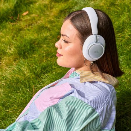 Una mujer de perfil descansando sobre el pasto al aire libre, vistiendo una camisa de rayas de colores y utilizando los audífonos circumaurales inalámbricos JBL Live 780NC de color blanco.