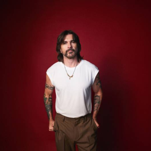 Fotografía de una persona de pie frente a un fondo rojo, con cabello oscuro a los hombros, barba, camiseta sin mangas blanca y brazos tatuados, mirando directamente a la cámara.