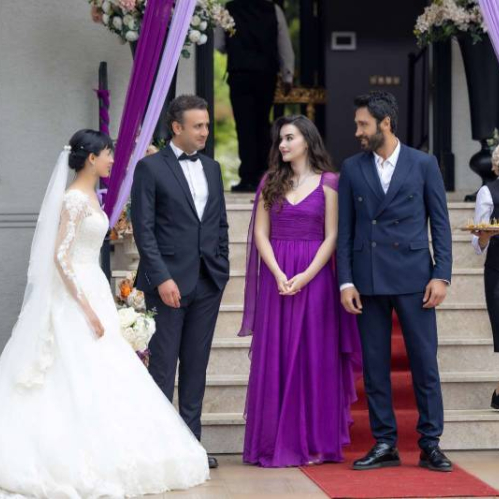 Fotografía de una ceremonia con cinco personas en un entorno decorado con flores, telas y escalinatas, donde una pareja vestida de gala interactúa con otros invitados mientras una mesera desciende con una bandeja de bebidas.
