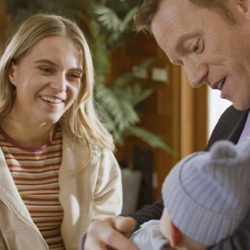 Fotografía promocional en plano medio de la película "Una Familia Perfecta" de Lifetime Movies. A la izquierda, una mujer joven rubia sonríe con calidez vistiendo una sudadera clara y playera a rayas. A la derecha, un hombre sonriente sostiene cuidadosamente en brazos a un bebé recién nacido envuelto con un gorrito tejido.