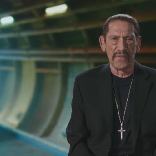 El actor Danny Trejo mira fijamente a la cámara en una toma de plano medio. Viste un saco negro, camiseta negra y un collar con un dije en forma de cruz plateada. Se encuentra posando en un estudio con un fondo abstracto de tonos azules que simula la estructura curva de un túnel o instalación industrial subterránea.