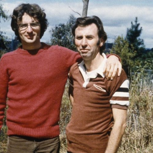 Fotografía de archivo en plano medio de dos hombres al aire libre. A la izquierda, un joven David Koresh con cabello castaño ondulado, lentes de armazón delgado y suéter de lana rojo abraza por el hombro a otro hombre a la derecha, quien viste una playera tipo polo marrón con rayas en las mangas. Ambos se encuentran frente a un terreno arbolado.