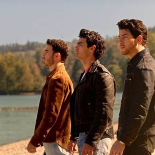 Fotografía en plano medio de perfil de los tres integrantes de los Jonas Brothers (Kevin, Joe y Nick Jonas) al aire libre frente a un cuerpo de agua con vegetación al fondo. Los tres miran hacia el horizonte de derecha a izquierda vestida cada uno con chaquetas casuales de tonos oscuro, café y cuero.