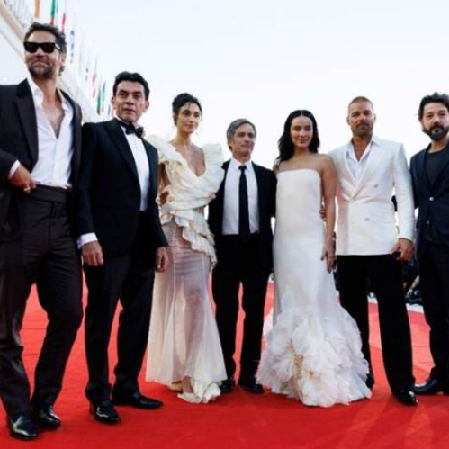Elenco y equipo de producción de la película "Hombre al Agua" posando sobre la alfombra roja del Festival Internacional de Cine de Venecia. De izquierda a derecha aparecen Manuel García-Rulfo, Jorge Salinas, Débora Nascimento, Gael García Bernal, Esmeralda Pimentel, Ricky Martin y Diego Luna vestida la mayoría en trajes de etiqueta y vestidos de gala en tonos blanco y negro.
