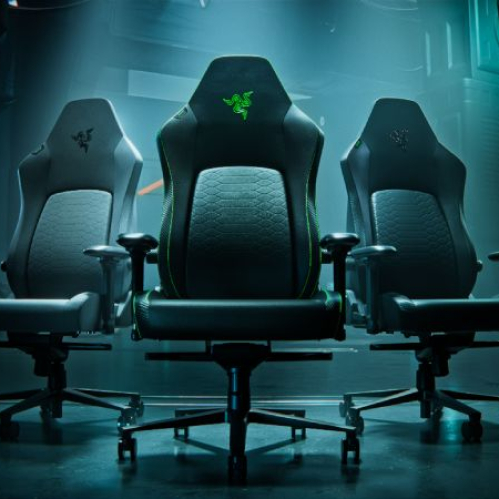Tres sillas gamer de diseño ergonómico en un entorno oscuro con iluminación tenue, destacando el logotipo verde de Razer en el reposacabezas.