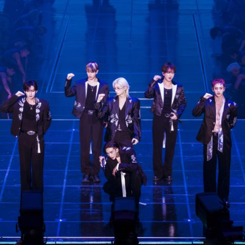 Fotografía del concierto de Stray Kids durante su gira mundial 'RUN IT' sobre un escenario principal iluminado por luces LED azules y neón. Los integrantes de la agrupación surcoreana lucen trajes oscuros coordinados con bordados en tono blanco de estilo tradicional. Siete de ellos están de pie realizando una pose coordinada con el brazo flexionado a la altura de la cabeza, mientras que Hyunjin aparece agachado en el centro del escenario frente al público.   