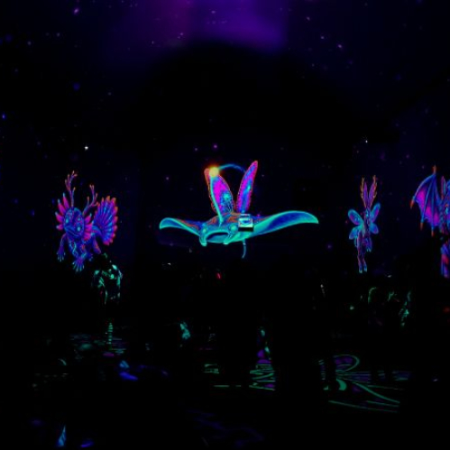 Proyección inmersiva en un espacio oscuro donde destacan alebrijes y figuras mitológicas voladoras iluminadas en tonos fluorescentes neón como morado, azul, rosa y naranja. Entre las criaturas se distinguen un ave con alas desplegadas, una mantarraya brillante y un mamífero alado rodeados de patrones prehispánicos en un entorno envolvente.