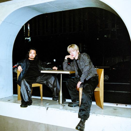 Dos personas sentadas en una estructura arquitectónica abierta hacia una calle nocturna, con estética urbana y tonos oscuros.