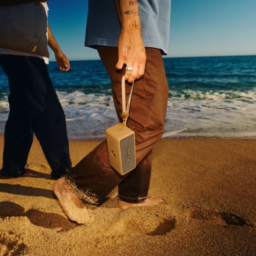 Una persona caminando descalza por la playa sosteniendo la bocina portátil Marshall Emberton III Brown mediante su correa, con el mar de fondo bajo la luz del sol.