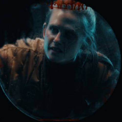 Fotografía de un personaje visto a través de una escotilla circular iluminada por un tono azulado, que refuerza la sensación de encierro y tensión en un entorno oscuro.