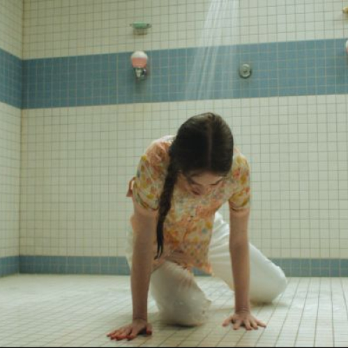 Fotografía de producción que muestra a una joven con vestido estampado de flores arrodillada en el piso de azulejos blancos de una regadera colectiva, con la cabeza inclinada bajo el chorro de agua que cae desde arriba.