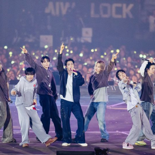 Integrantes de la agrupación de K-pop BTS actuando sobre el escenario de un estadio durante un concierto. Los siete miembros viste ropa casual de estilo urbano y mezclilla mientras interactúan con el público en un escenario iluminado, con espectadores al fondo sosteniendo luces de concierto.