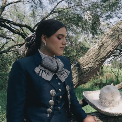 Camila Fernández luce un traje charro azul marino con moño gris bordado y detalles plateados mientras mira hacia abajo en un entorno natural con un sombrero charro blanco a su lado. Archivo: General 2.jpg.