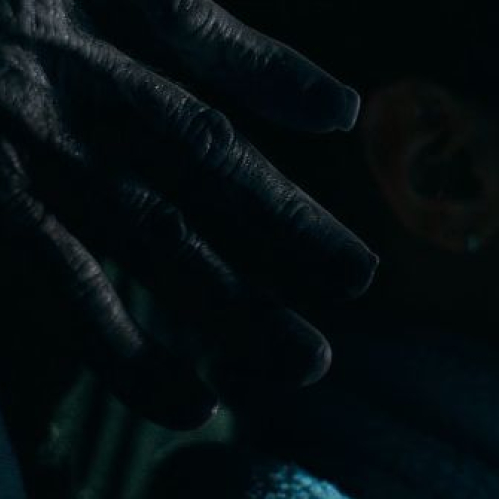 Mano oscura acercándose al hombro de una persona en una escena de tensión con iluminación tenue.