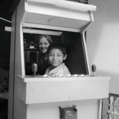 Fotografía en blanco y negro detrás de cámaras (BTS) en un set de filmación. En el centro, un niño de cabello corto y sonrisa leve se asoma desde la estructura de una máquina recreativa tipo arcade de madera. Detrás de la máquina de arcade se observa a una mujer sonriente con audífonos. A la izquierda aparece de perfil un miembro del equipo de producción junto a un pedestal con equipo técnico de iluminación y cámara.