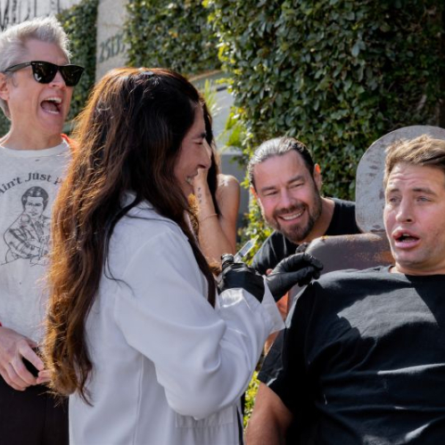 Fotografía promocional de Jackass: La Última y Nos Vamos donde Johnny Knoxville, vistiendo un suéter rojo y anteojos de sol, ríe efusivamente junto a Chris Pontius mientras una especialista médica aplica una inyección en el labio de un participante sentado en una silla.