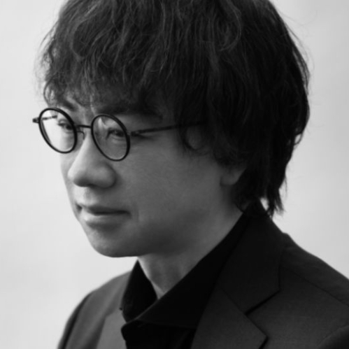 Retrato fotográfico en blanco y negro del director japonés Makoto Shinkai de perfil tres cuartos. El cineasta viste un saco oscuro sobre camisa negra y lleva anteojos de armazón redondo.