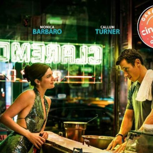 Fotografía en formato horizontal de una escena de la película dentro de un establecimiento nocturno en Nueva York. A la izquierda, la actriz Monica Barbaro luce un vestido verde brillante de lentejuelas con escote halter y sostiene una botella mientras apoya los brazos en la barra. Frente a ella, a la derecha, el actor Callum Turner la mira detenidamente mientras viste una playera tipo polo gris con una toalla blanca al hombro tras la barra. En el fondo se aprecia un letrero neón verde reflejado con la palabra "CLAREMONT" y en la esquina superior derecha se ubica el sello circular de "Garantía Cinépolis".