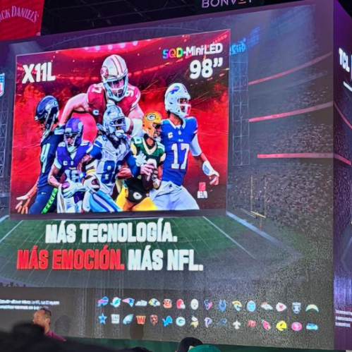 Una pantalla gigante cúbica en un evento de presentación donde se muestra la televisión TCL X11L de 98 pulgadas con tecnología SQD-MiniLED y gráficos de jugadores de la NFL sobre una cancha virtual.