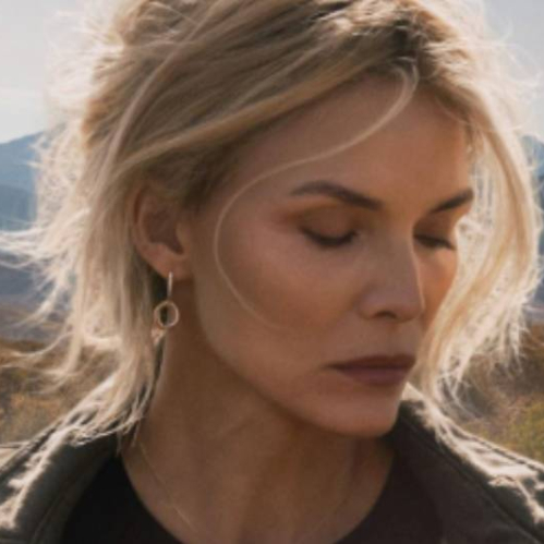 Persona de cabello claro recogido, con expresión introspectiva, situada frente a un paisaje de montañas y árboles otoñales iluminados por luz natural.