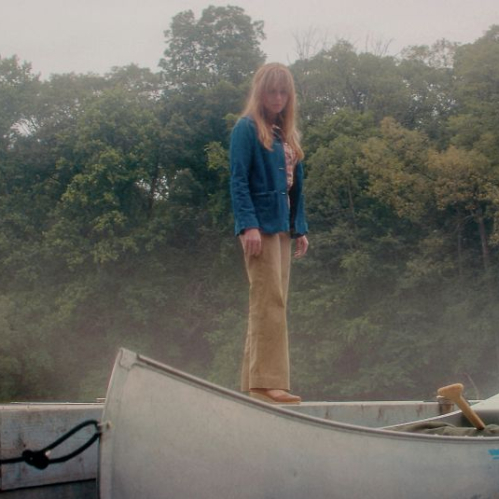 Persona de pie junto a una canoa en un muelle rodeado de niebla y vegetación, en un ambiente de tensión y misterio.