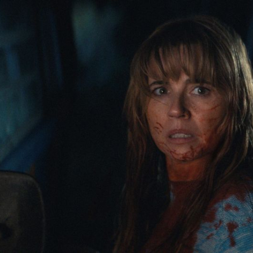 Fotograma de la serie Crystal Lake donde la actriz Linda Cardellini interpreta a Pam Voorhees. Aparece en plano medio con rostro aterrorizado, cabello largo castaño con fleco y salpicaduras de sangre en la mejilla y en su suéter azul con patrones blancos. De fondo se aprecia la noche oscura y el espejo retrovisor de un vehículo.