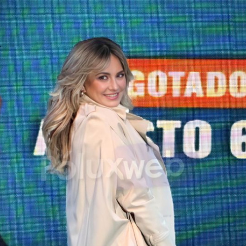 Fotografía de María José sonriendo de perfil luciendo un abrigo beige elegante frente a una pantalla LED de eventos que muestra el letrero "AGOTADOS" en letras rojas y amarillas.