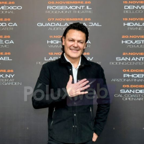 Fotografía en plano medio de Pedro Fernández sonriendo con una mano sobre el pecho y vistiendo chamarra negra y camisa blanca, posando frente a un banner promocional gris con las fechas y ciudades de la gira "Ave Fénix Two'r 2026".