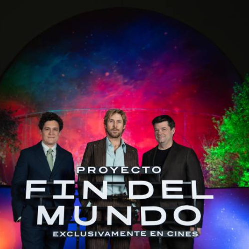 Tres integrantes del equipo de la película posan frente a una estructura iluminada con colores vibrantes, acompañados por un letrero promocional del filme en un entorno nocturno.