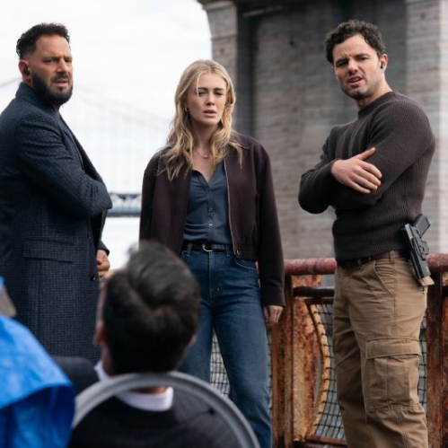Fotografía de una escena exterior de la segunda temporada de la serie The Hunting Party. Tres investigadores permanecen de pie junto a un barandal de metal oxidado cerca de un río, con un gran puente de suspensión y un muelle de concreto de fondo bajo un cielo nublado. A la izquierda, un hombre con barba y abrigo gris oscuro mira con desconcierto hacia abajo. En el centro, una mujer rubia con chaqueta marrón y jeans muestra preocupación. A la derecha, un hombre con suéter café y pantalón táctico beige tiene los brazos cruzados y una pistola en su funda al cinto. En primer plano, de espaldas, se observa parcialmente la cabeza de una persona en una silla y una lona azul.