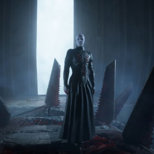 Captura de pantalla en alta definición del videojuego de terror "Clive Barker’s Hellraiser: Revival". En el centro de la escena, el icónico personaje Pinhead posa de pie vistiendo su clásica túnica larga de cuero negro con detalles de heridas abiertas en el pecho. Se encuentra en medio de una estructura gótica o templo oscuro dentro del Laberinto, rodeado por gigantescas hojas de sierra circulares oxidadas y ensangrentadas que sobresalen del suelo. Al fondo, una intensa luz blanca y azulada resalta la silueta del personaje, creando una atmósfera de suspenso y neblina.