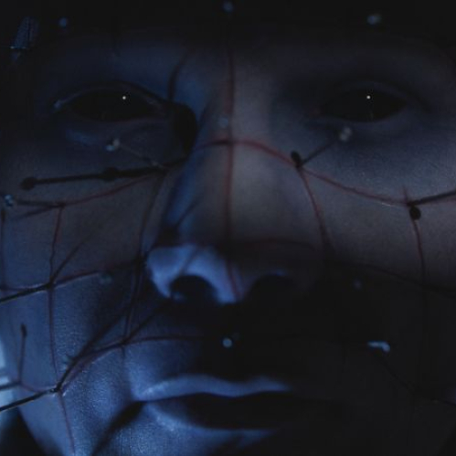Primer plano de un rostro iluminado en tonos azules, atravesado por múltiples clavos metálicos conectados por líneas rojas, evocando una estética oscura y perturbadora.