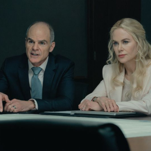 Fotografía de producción de la serie Lioness donde se observa a Michael Kelly y Nicole Kidman sentados detrás de una mesa de juntas de madera. Michael Kelly, del lado izquierdo, viste un traje oscuro con camisa blanca y corbata azul texturizada mientras gesticula y señala hacia la mesa con su mano derecha. Nicole Kidman, del lado derecho, viste un saco formal de color rosa pálido, mantiene sus manos entrelazadas sobre la mesa y observa fijamente hacia el frente con una expresión seria.