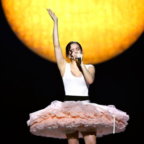 Artista sobre el escenario interpretando frente a un fondo circular luminoso, con vestuario escénico en tonos claros y falda amplia, capturada en un momento de energía y expresión durante su presentación.