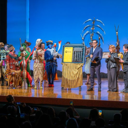 Fotografía del elenco del musical El Rey León en el escenario junto a ejecutivos y creativos durante la develación de la placa conmemorativa. Al centro se observa la placa colocada sobre un atril dorado mientras los intérpretes caracterizados con sus vestuarios y máscaras aplauden.