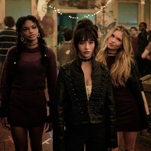 Tres jóvenes actrices interpretando a brujas luciendo ropa oscura y accesorios de época mientras caminan juntas en una fiesta escolar iluminada con luces cálidas.