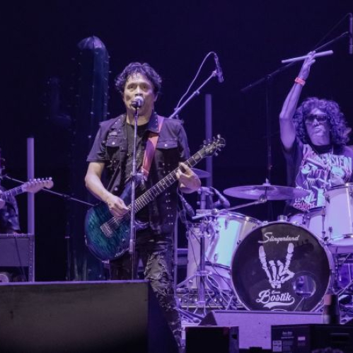 Banda de rock interpretando en vivo con guitarras eléctricas, batería y luces púrpura.