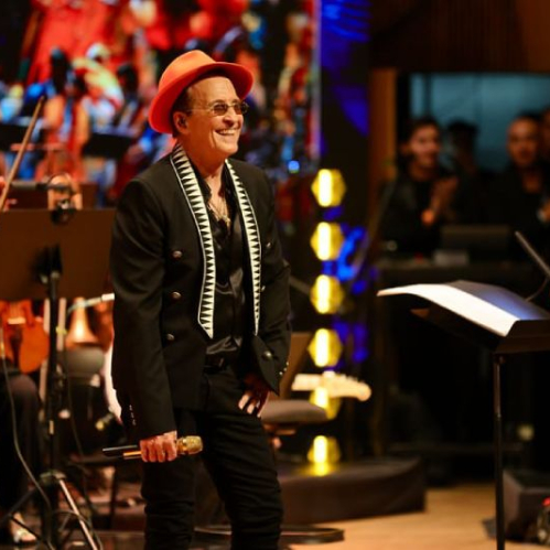 Fotografía en vivo del cantante Emmanuel sonriendo en el escenario con sombrero naranja y saco negro, micrófono en mano, rodeado por los músicos de una orquesta sinfónica con sus instrumentos de cuerda.