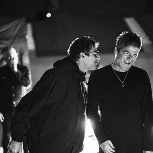 Fotografía en blanco y negro de los hermanos Liam y Noel Gallagher en el área de vestidores durante su gira. A la izquierda, Liam viste una chamarra oscura con gorro y anteojos de sol mientras le habla al oído a Noel. A la derecha, Noel sonríe vistiendo una playera oscura de manga larga con una cadena al cuello.