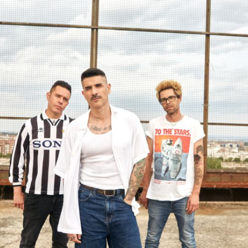 Los tres integrantes de la banda de rock Pignoise posan al aire libre sobre una azotea urbana frente a una malla metálica. El vocalista viste playera blanca abierta y jeans, flanqueado por sus compañeros con playeras estampadas.   
