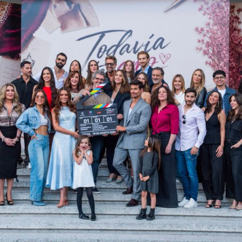 Fotografía grupal al aire libre en unas escalinatas de piedra con el elenco completo y equipo de producción de la telenovela Todavía te amo. En el centro, los protagonistas y productores sostienen una claqueta de producción con el logotipo del melodrama contra un fondo promocional con temática de sastrería y costura.